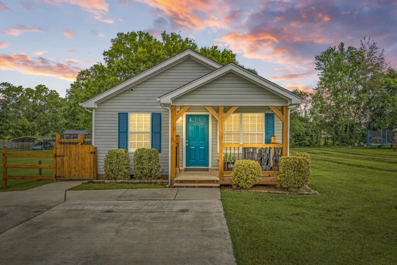Front exterior of a cottage-style home with a teal door and covered porch, photographed at dusk under a pink sky
