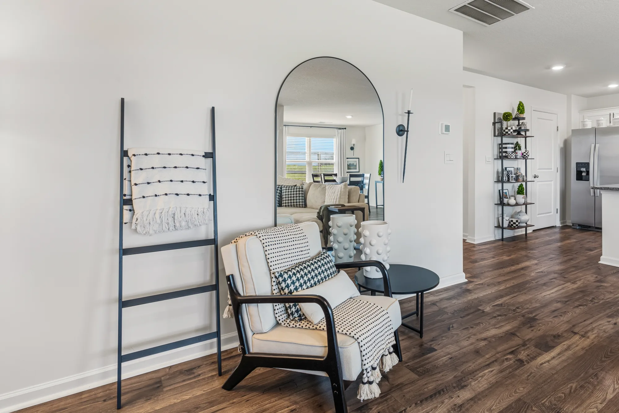 Bright staged living area with an arched mirror, black accent chair, and an open sightline through to the kitchen
