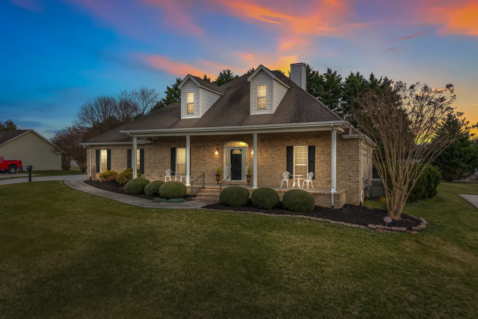 Brick home with a covered porch and dormer windows at twilight, windows lit warm against a pink and blue sky