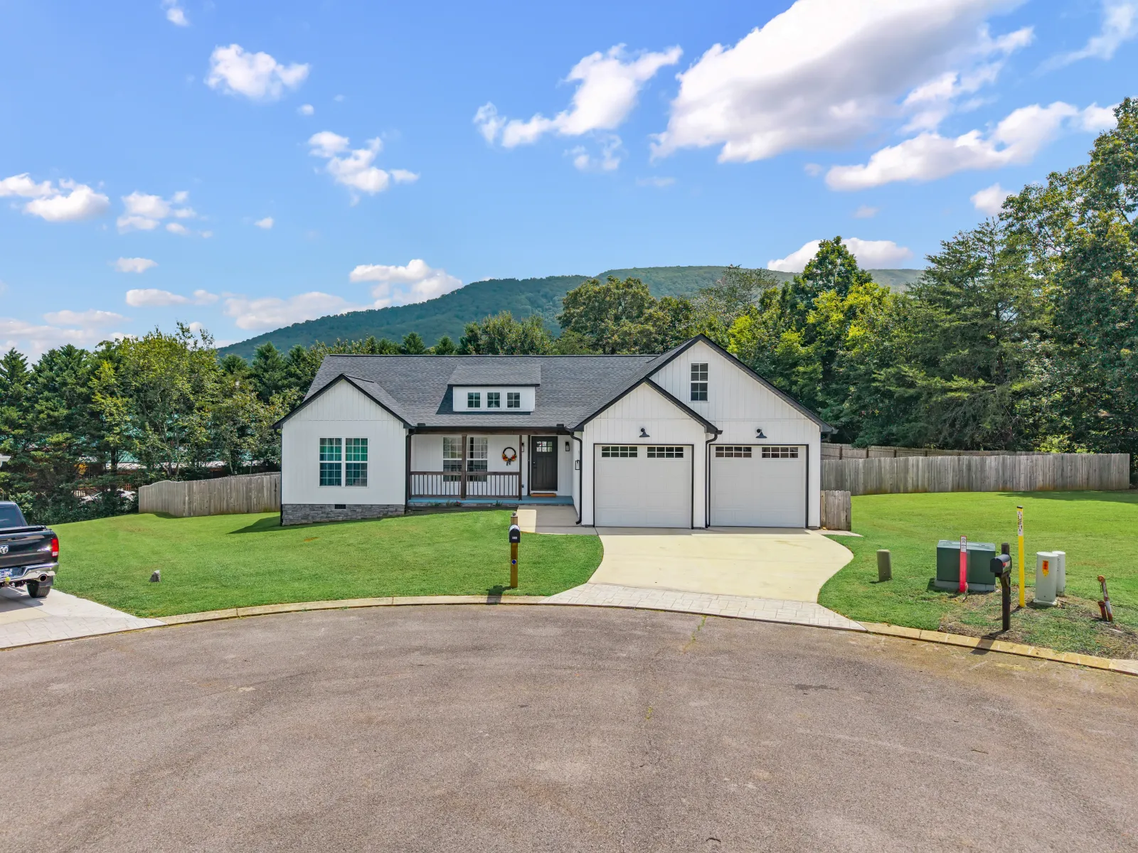 White farmhouse-style home with a two-car garage at the end of a cul-de-sac, a wooded ridge rising behind it